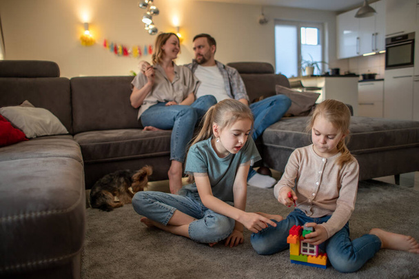 дети, сестры весело играют дома на полу, а родители отдыхают на диване - Фото, изображение