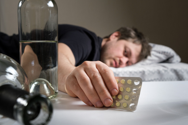 A man lies on a bed after an alcohol party, holding hangover pills in his hand - Photo, Image