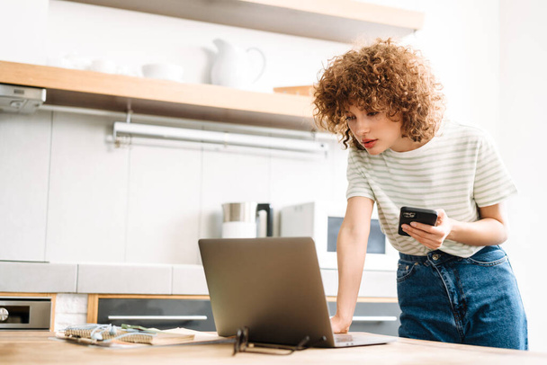 European woman working with laptop and cellphone while standing at home kitchen - Foto, Imagen