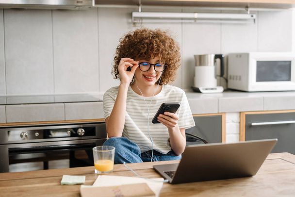 Joven estudiante feliz mujer en auriculares usando el teléfono celular mientras estudia con el ordenador portátil en la cocina del hogar - Foto, Imagen