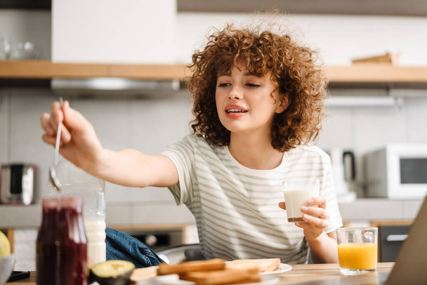 Smiling young woman having breakfast while sitting at table in home kitchen - Foto, Bild
