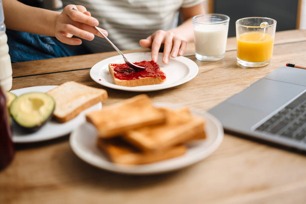 Jeune femme européenne utilisant un ordinateur portable tout en prenant le petit déjeuner à la maison cuisine - Photo, image
