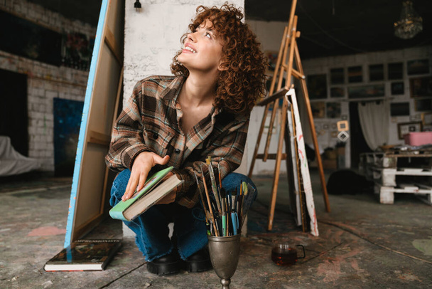 Smiling young white redhead woman sitting in an art studio next to canvas holding book - Фото, изображение