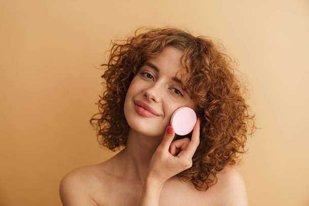 Half-naked ginger woman smiling while showing cosmetic sponge isolated over beige background - Fotó, kép