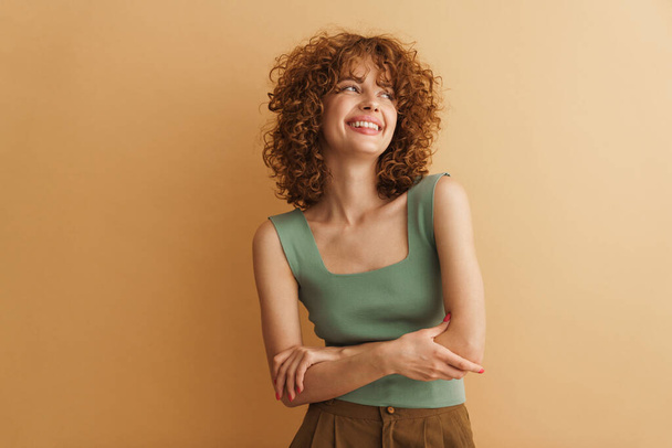 Smiling young casual woman with curly hair standing looking aside over beige background - Valokuva, kuva