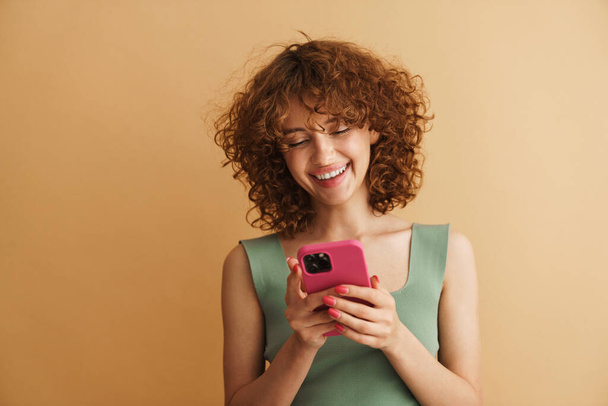 Bej arka planda izole edilmiş cep telefonuyla gülen genç bir kızıl kadın. - Fotoğraf, Görsel