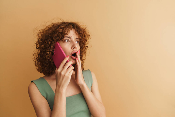 Excited shocked young casual woman with curly hair talking on mobile phone standing over beige background - Photo, image
