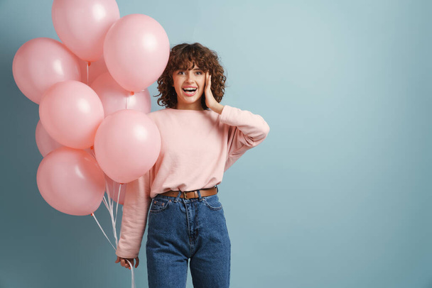 Young ginger happy woman smiling while posing with pink balloons isolated over blue background - Valokuva, kuva