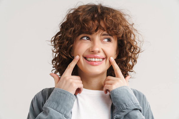 Ginger joyful woman smiling and pointing fingers at her cheeks isolated over white background - Foto, Bild