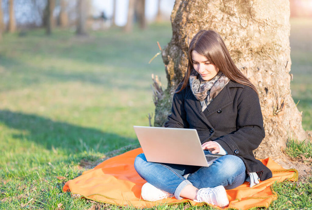 公園の木のふもとに座ってノートパソコンで働く美しい若いブルネットの女性。彼女はキーボードをタイプする。リモートワークの概念。スペースのコピー. - 写真・画像
