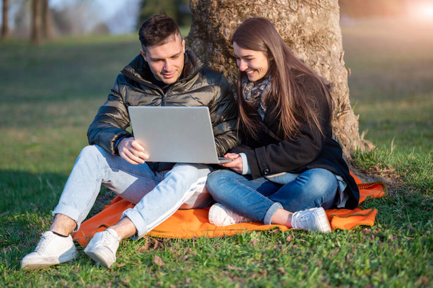 A beautiful couple in casual wear is having fun with the laptop outdoors. Sitting at the foot of a tree, you enjoy nature on a sunny spring day. Remote teamwork concept. - Fotó, kép