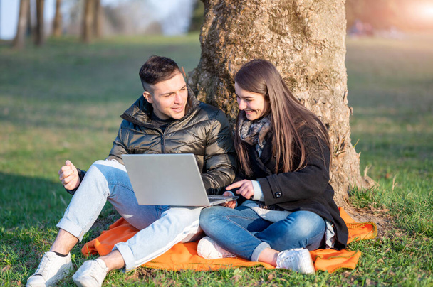 Un beau couple en tenue décontractée s'amuse avec l'ordinateur portable à l'extérieur. Assis au pied d'un arbre, vous profitez de la nature par une journée de printemps ensoleillée. Concept de travail d'équipe à distance. - Photo, image