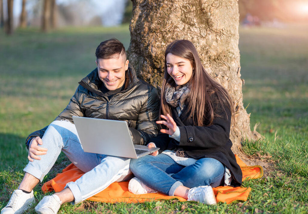 A beautiful couple in casual wear is having fun with the laptop outdoors. Sitting at the foot of a tree, you enjoy nature on a sunny spring day. Remote teamwork concept. - Photo, Image