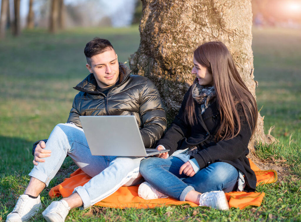 A beautiful couple in casual wear is having fun with the laptop outdoors. Sitting at the foot of a tree, you enjoy nature on a sunny spring day. Remote teamwork concept. - Foto, afbeelding