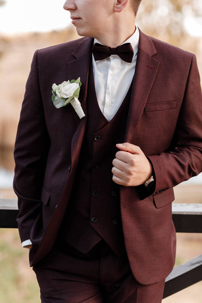 the groom in a bordo wedding suit, a tuxedo in a white shirt, bow tie and a roses buttonhole. High quality photo. - Zdjęcie, obraz
