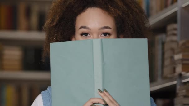 Közelkép fiatal boldog játékos lány diák bujkál könyv mögött kukucskál ki a tankönyv mosolygós fogas mosoly örül játszik flörtölés nevetés nézi kamera élvezi olvasás egyetemi könyvtár - Felvétel, videó