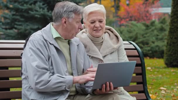 Happy carefree elderly senior grandparents sit on bench in autumn park man typing on laptop together reading discussing internet news mature couple make online order easy use modern technology concept - Footage, Video
