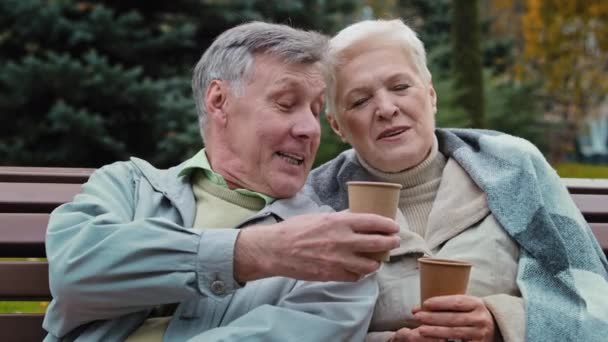 Счастливая пожилая пара разговаривает сидеть в осеннем парке отдохнуть вместе беззаботно пожилые бабушка и дедушка пить кофе чай на открытом воздухе наслаждаясь приятной беседой улыбается женщина мужчина говорит расслабляющий на скамейке - Кадры, видео
