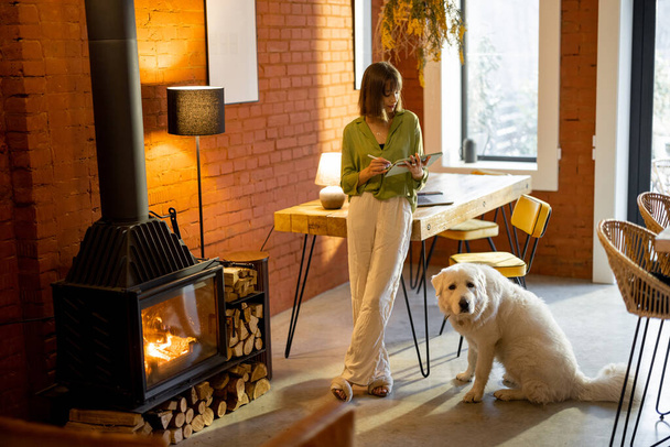 Γυναίκα εργάζεται σε ψηφιακή ταμπλέτα, ενώ στέκεται με το σκύλο της στο άνετο σαλόνι - Φωτογραφία, εικόνα
