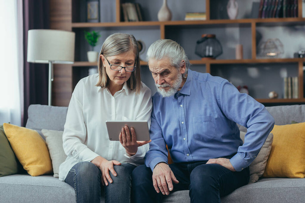 Starsza siwowłosa para, mężczyzna i kobieta, siedzący na kanapie w domu. korzystanie z komputera typu tablet do komunikacji wideo i konsultacji online z lekarzem - Zdjęcie, obraz