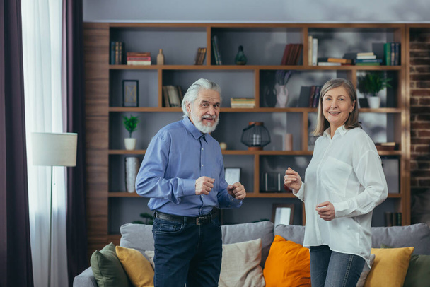 Happy cheerful senior married couple having fun at home. older family is dancing. Gray haired retirees rejoice moving active lifestyle. Old man and woman spend leisure time indoors listen to music - Фото, зображення