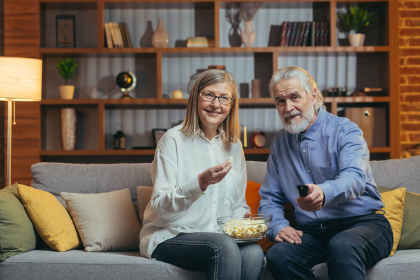 grey-haired older couple at home choose a television show to watch and switch channels with a TV remote control. elderly mature family husband and wife cannot choose a show. watching news or film - Fotografie, Obrázek