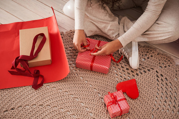 Vielrassige Frau wickelt Geschenk mit rotem Papier ein und dekoriert es mit Schleife in ihrem Zimmer am Boden - Foto, Bild