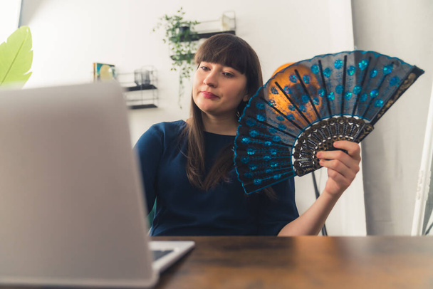 Pretty young dark haired woman enjoys working from home, looking at the screen, having a folding fan in her hand living room background remote job concept copy space - Foto, Bild