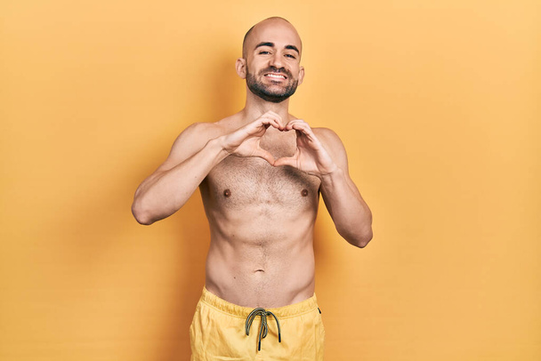 Young bald man wearing swimwear smiling in love doing heart symbol shape with hands. romantic concept.  - Photo, Image