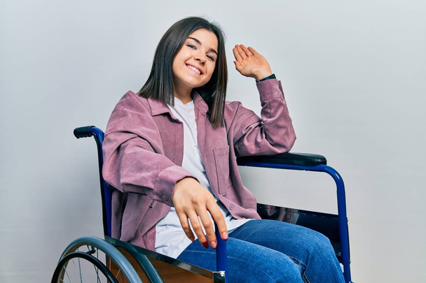 Młoda brunetka siedzi na wózku inwalidzkim rezygnując mówiąc cześć szczęśliwy i uśmiechnięty, przyjazny gest powitalny  - Zdjęcie, obraz