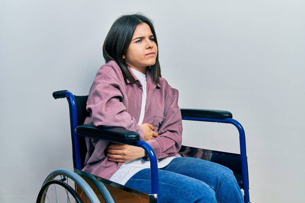 Junge brünette Frau sitzt im Rollstuhl mit der Hand auf dem Bauch, weil Verdauungsstörungen, schmerzhafte Krankheit, die sich unwohl fühlt. Schmerzkonzept.  - Foto, Bild