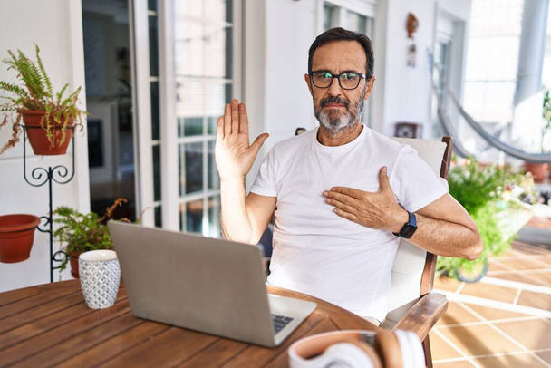 Mann mittleren Alters mit Computer-Laptop zu Hause fluchend mit der Hand auf der Brust und offener Handfläche, Treueschwur  - Foto, Bild