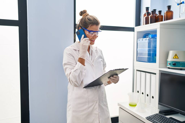 jong blond vrouw dragen wetenschapper uniform praten op de smartphone in laboratorium - Foto, afbeelding