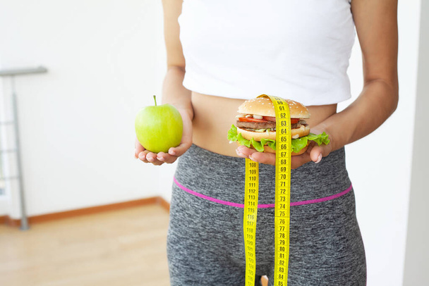 Giovane donna in possesso di hamburger grasso dannoso con nastro di misurazione giallo - Foto, immagini