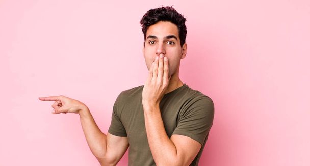 handsome hispanic man feeling happy, shocked and surprised, covering mouth with hand and pointing to lateral copy space - Foto, immagini