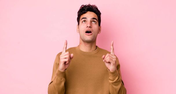 handsome hispanic man looking shocked, amazed and open mouthed, pointing upwards with both hands to copy space - Foto, Bild