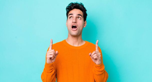 handsome hispanic man looking shocked, amazed and open mouthed, pointing upwards with both hands to copy space - Foto, imagen