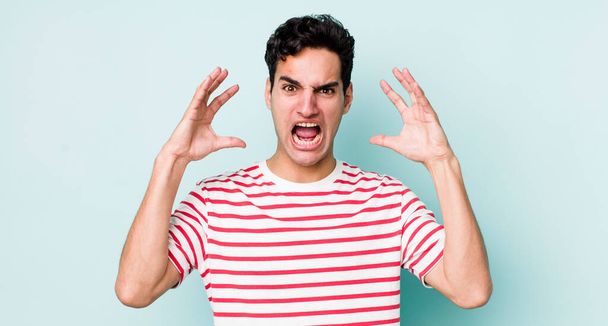 handsome hispanic man screaming with hands up in the air, feeling furious, frustrated, stressed and upset - Foto, afbeelding