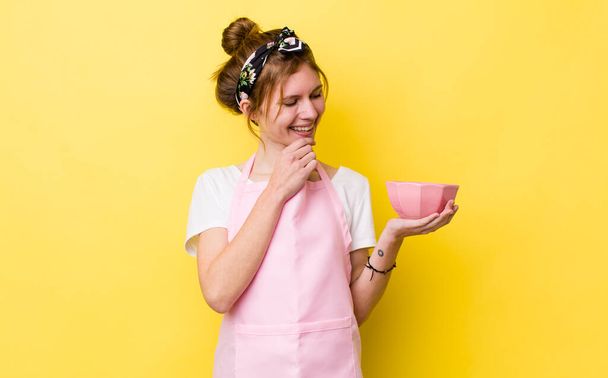 red head pretty woman smiling with a happy, confident expression with hand on chin. housewife holding an empty bowl - Foto, Imagen