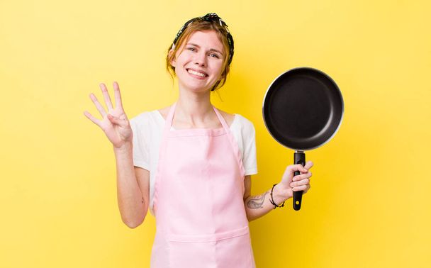 red head pretty woman smiling and looking friendly, showing number four. fry pan and housewife concept - Photo, Image