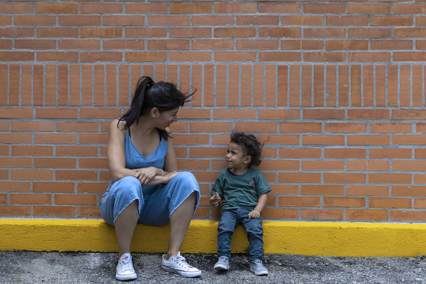 Latin American Mom chats with her son while they sit down to rest, brick wall background. Motherhood and Mother's Day concept. - Фото, изображение