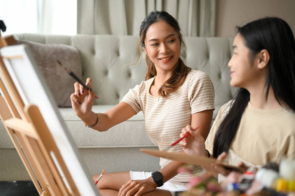 Krásná asijská sestra učí svou mladší sestru malovat akrylovou barvu na plátno, dělat kreativní koníčky společně v jejich domě. - Fotografie, Obrázek