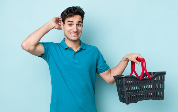 handsome hispanic man feeling stressed, anxious or scared, with hands on head. shopping basket concept - 写真・画像