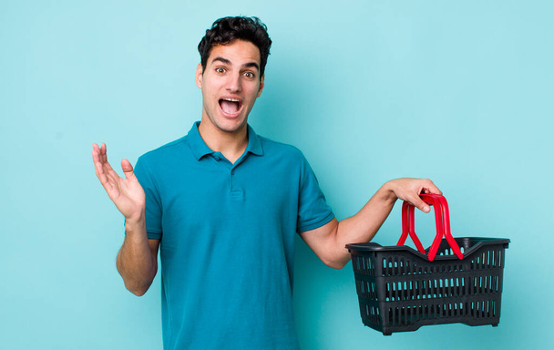 handsome hispanic man feeling happy and astonished at something unbelievable. shopping basket concept - Фото, изображение