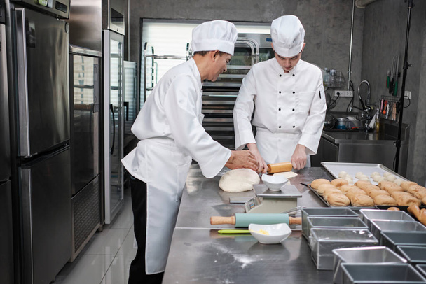 Two professional Asian male chefs in white cook uniforms and aprons are kneading pastry dough and eggs, preparing bread and fresh bakery food, baking in oven at stainless steel kitchen of restaurant. - 写真・画像