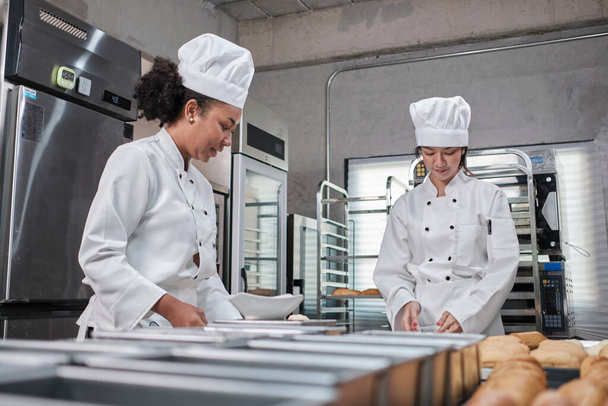 Two professional female chefs in white cook uniforms and aprons knead pastry dough and eggs, prepare bread, cookies, and fresh bakery food, baking in oven at a stainless steel kitchen of a restaurant. - Photo, Image