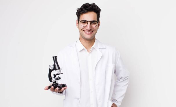 handsome hispanic man smiling happily with a hand on hip and confident.. scientist with microscope concept - Foto, immagini