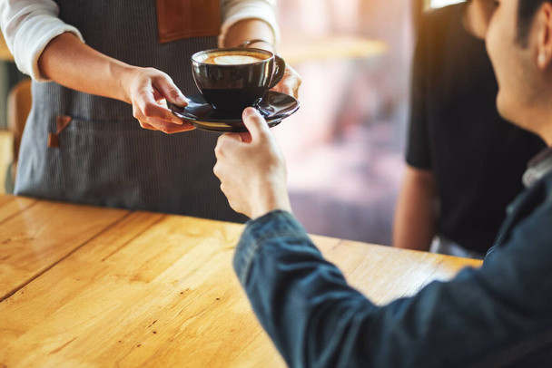Servírka drží a podává šálek horké kávy mužskému zákazníkovi v kavárně - Fotografie, Obrázek