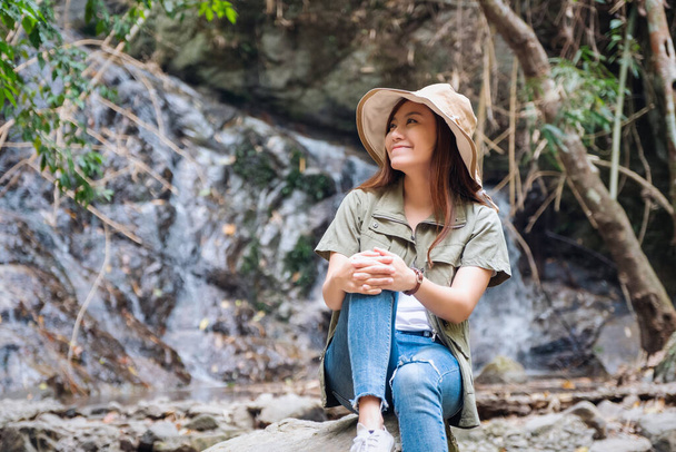 滝の前の岩の上に座っている美しいアジアの女性 - 写真・画像