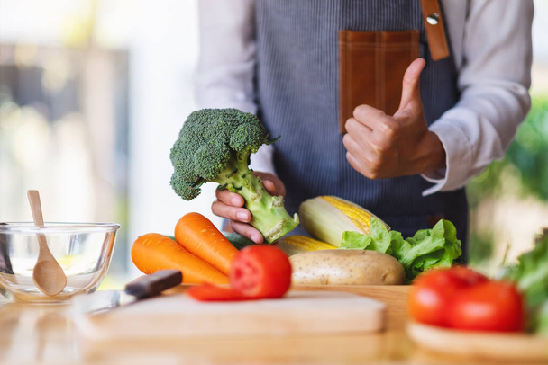 Kobieta szef kuchni trzyma kciuk w górze, aby pokazać dobry znak zielonym brokułom z tacą warzywną na stole - Zdjęcie, obraz